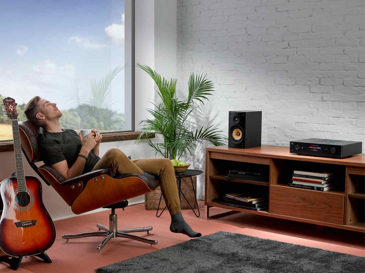 Cozy modern living room with a person sitting in a brown leather lounge chair holding a mug beside an acoustic guitar and a wooden media console with speakers and records.