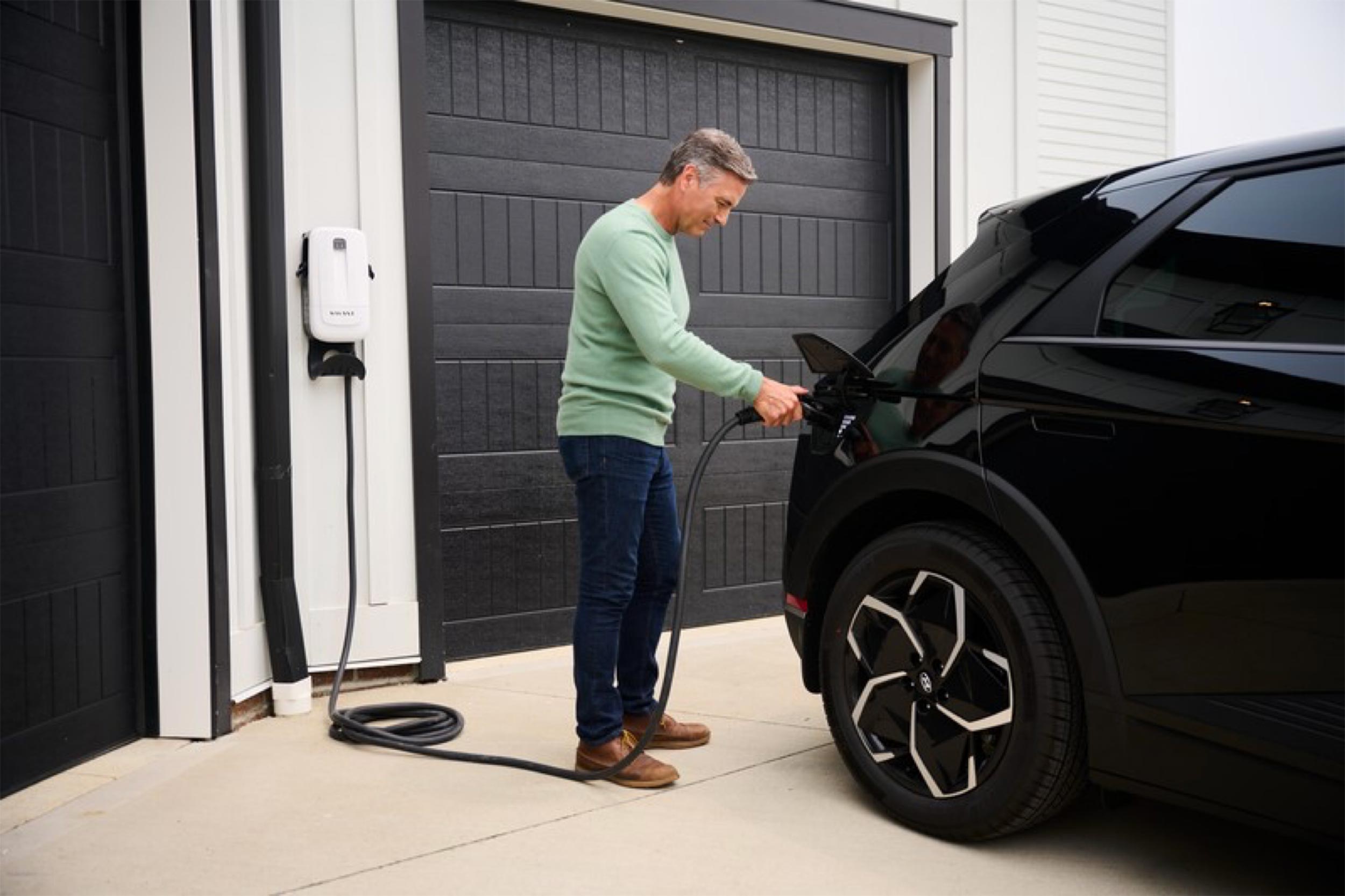 Man in green sweater charging a black electric vehicle in a driveway; cable runs from a wall-mounted white home charger beside a black garage door.