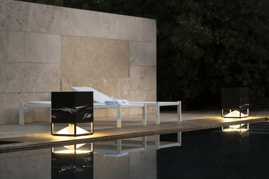 Lanterns light up two lounge chairs poolside near a stone wall.