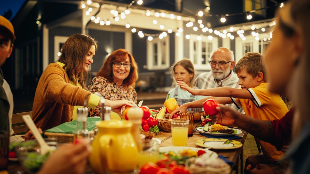Happy family and friends gather around an outdoor dinner table under string lights, sharing food and conversation in a cozy backyard setting.