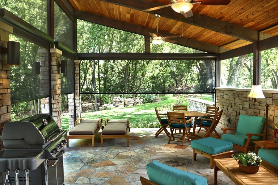 Retractable shades halfway lowered around a patio with a grill, tables and chairs, and a wood ceiling with fans. 
