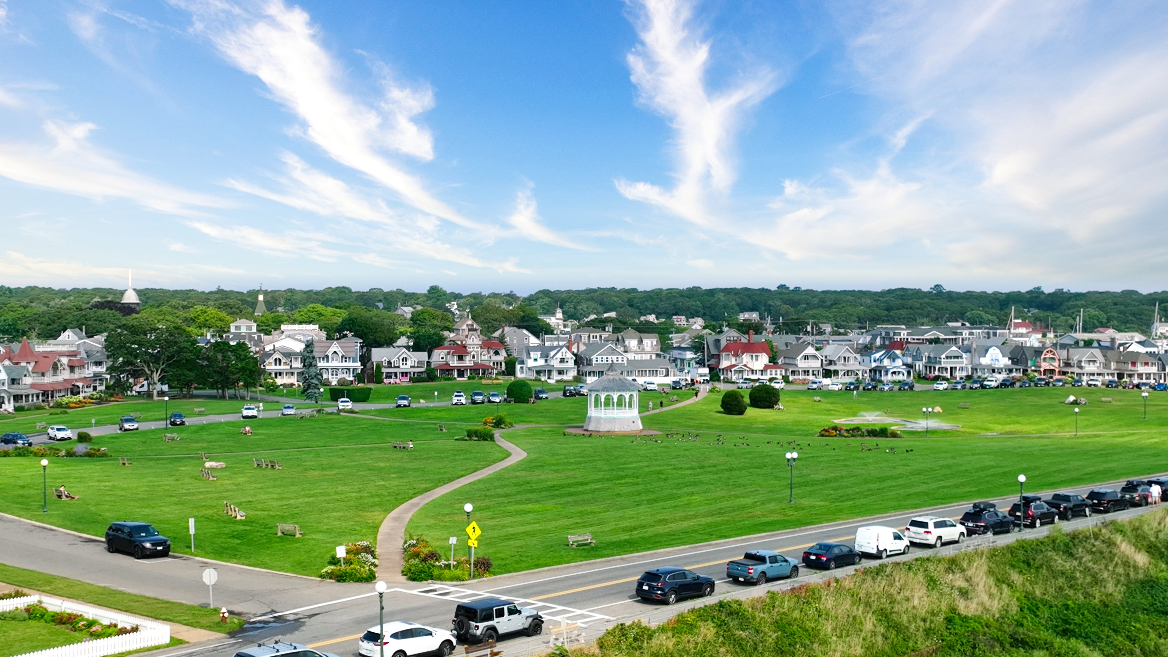 Wide view of a coastal New England town with a large green park and white gazebo in the center, lined by Victorian-style houses and a road filled with parked cars under a bright blue sky with wispy clouds.