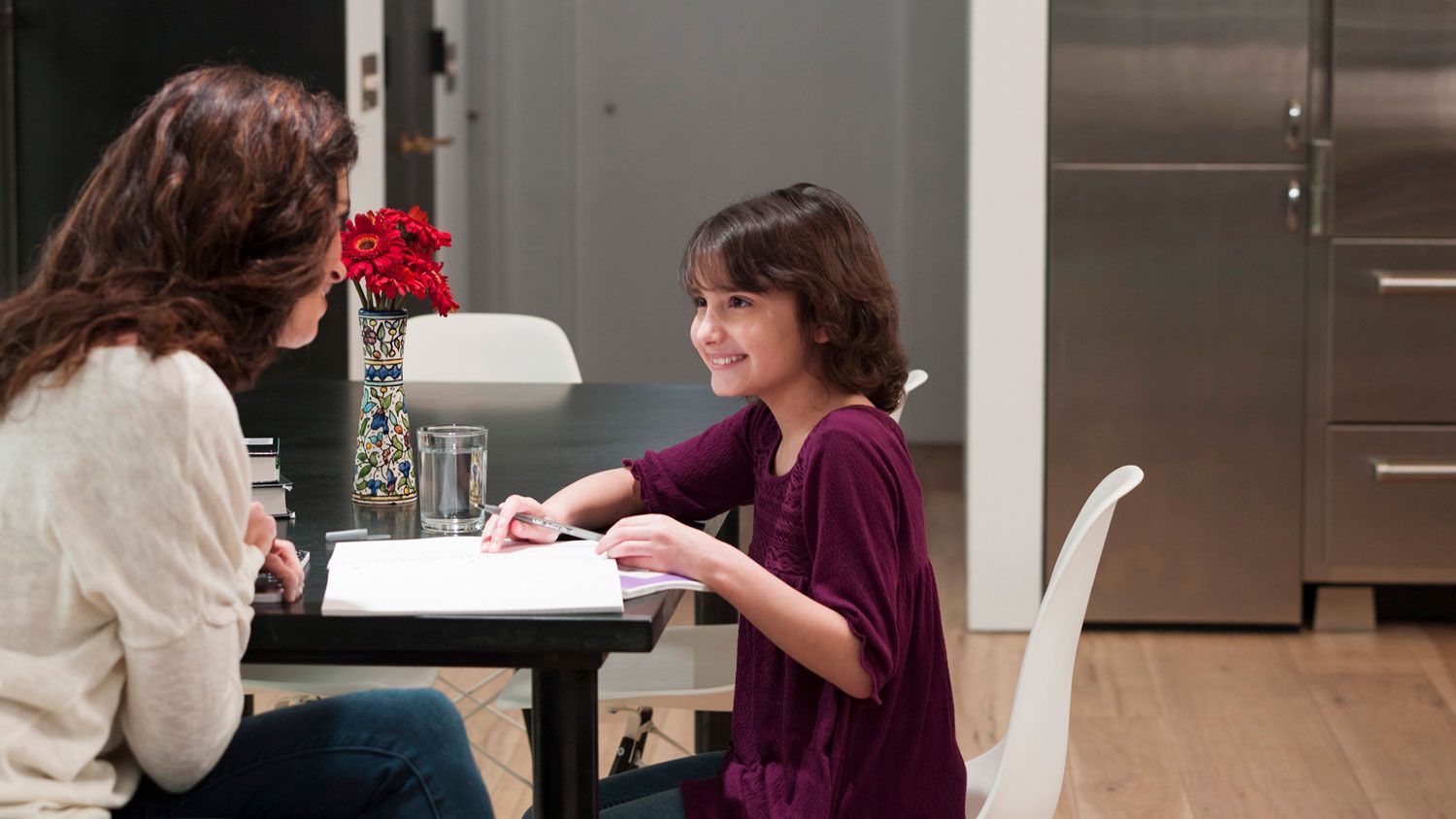 Smiling young girl sitting at a dining table doing homework with a woman across from her, in a modern kitchen with light wood floors and a vase of red flowers on the table.